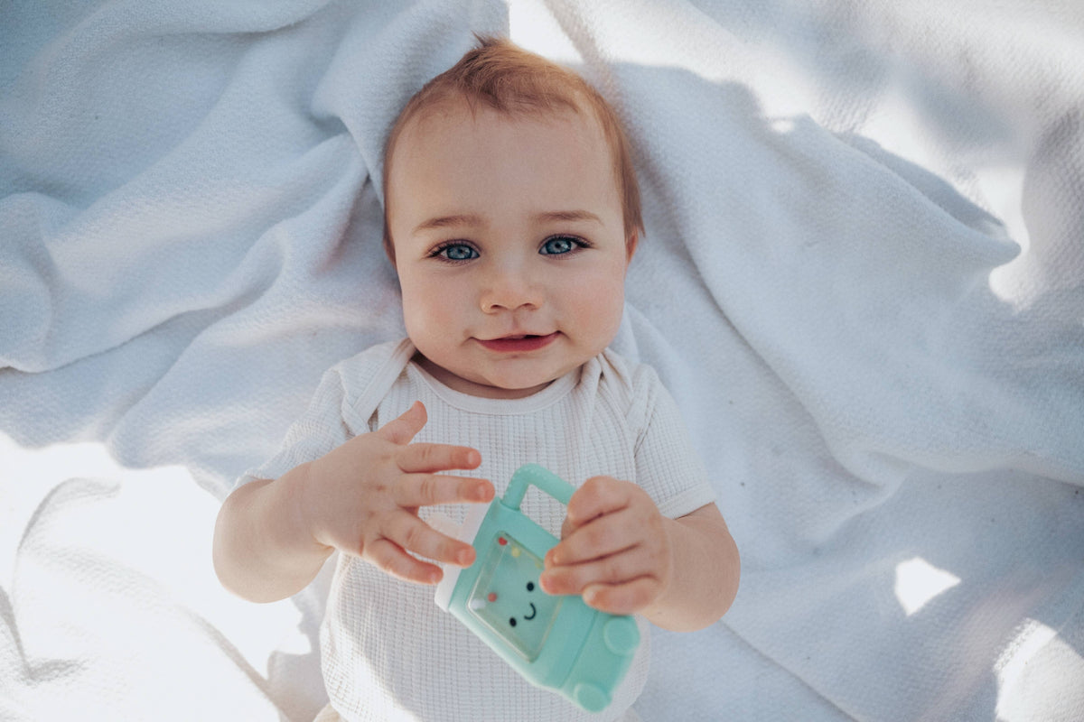 Baby holding a green teether on a white blanket