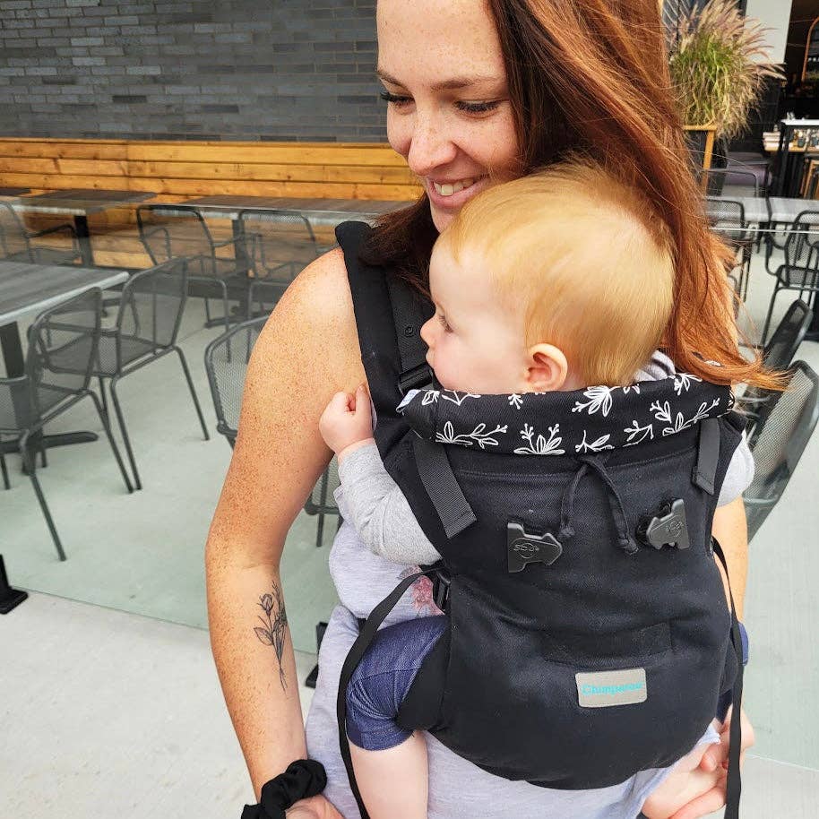Woman carrying a baby in a black carrier with floral patterns in an outdoor setting.